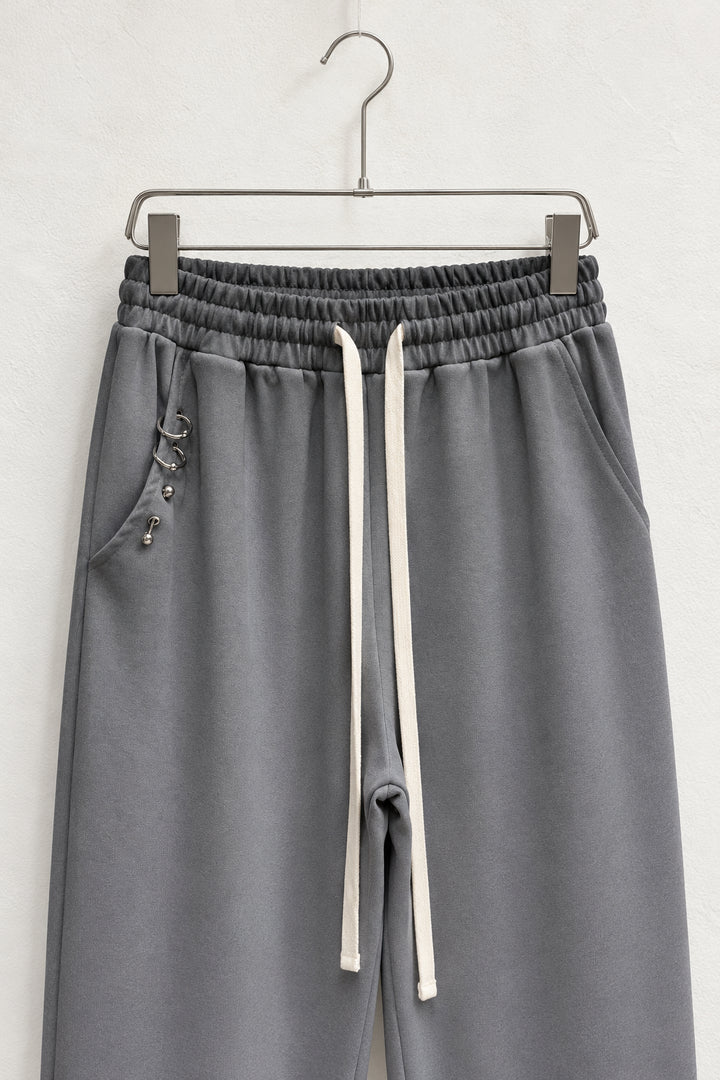 Detaliu talie elastică, șnururi plate și buzunar cu inele metalice la pantalonii gri.