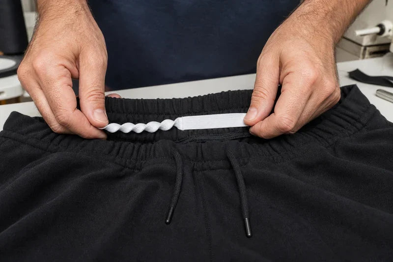 Mâini ținând o bandă elastică albă răsucită deasupra beteliei unor pantaloni negri cu două șnururi negre