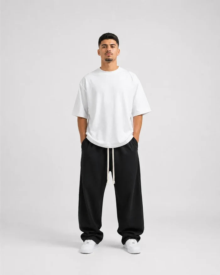 Bărbat în pantaloni negri largi cu șnur alb lung și tricou alb, pe fundal deschis