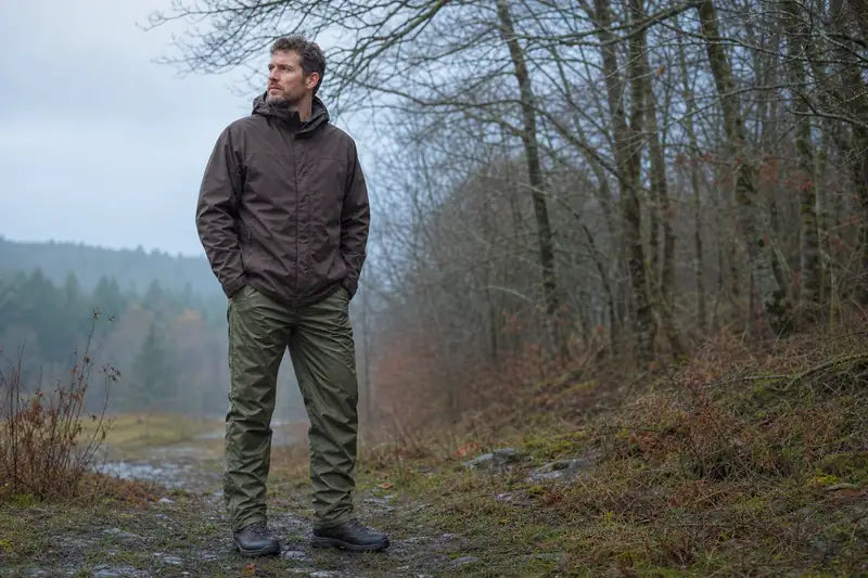 Bărbat în pantaloni olive și jachetă maro pe o potecă forestieră umedă