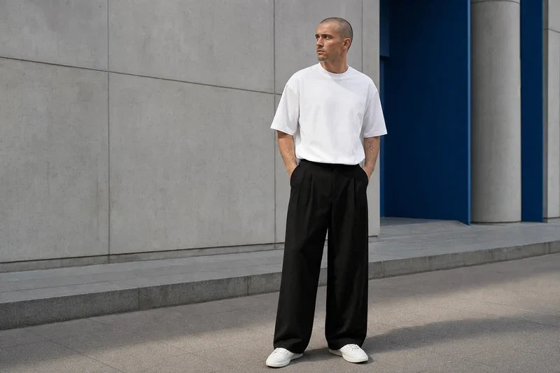 Bărbat adult în pantaloni wide leg negri, tricou alb și sneakers albi, lângă o clădire