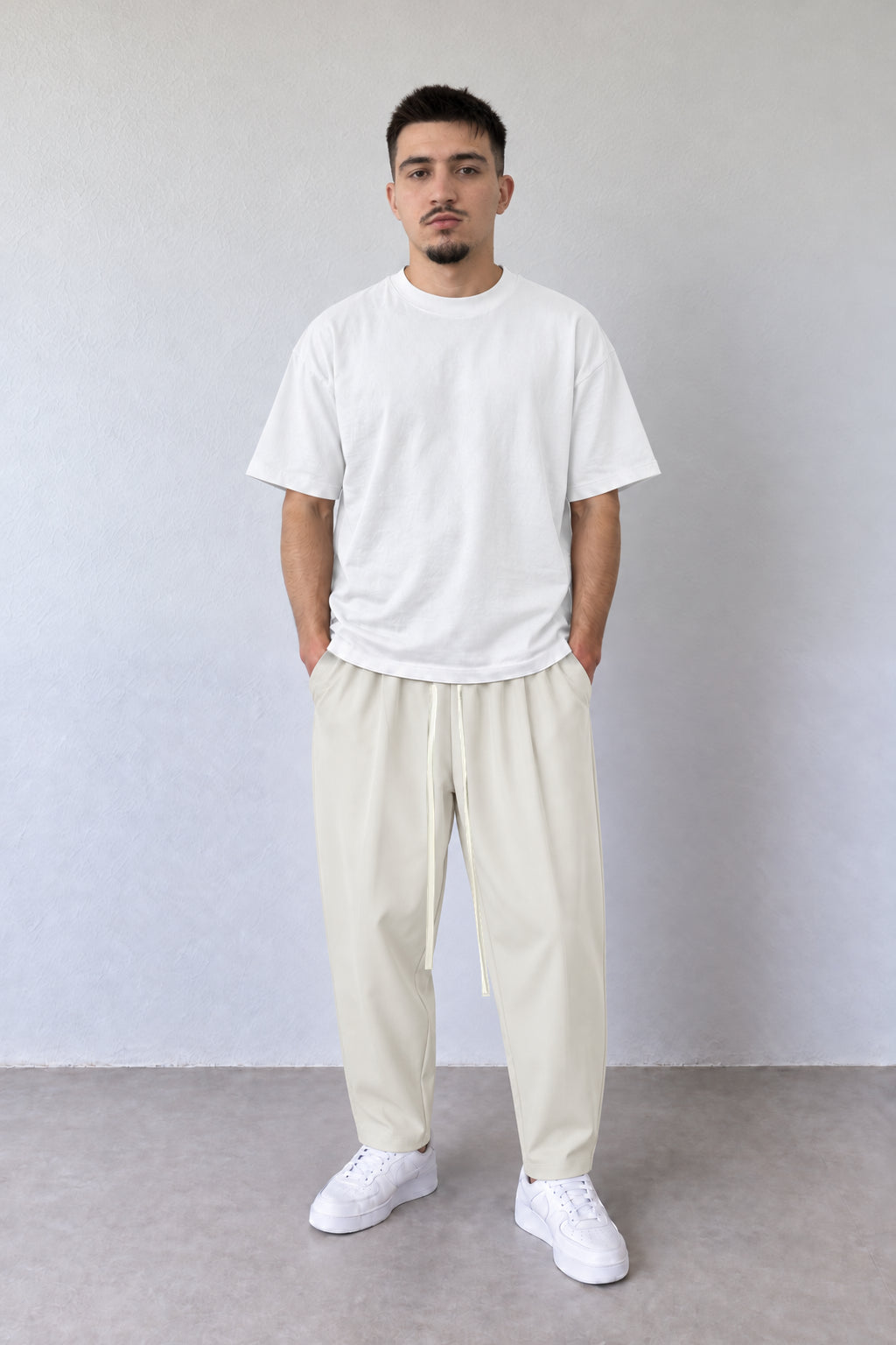 Pantaloni crem carrot fit pentru bărbați, cu șnur lung, purtați cu tricou alb, vedere integrală.