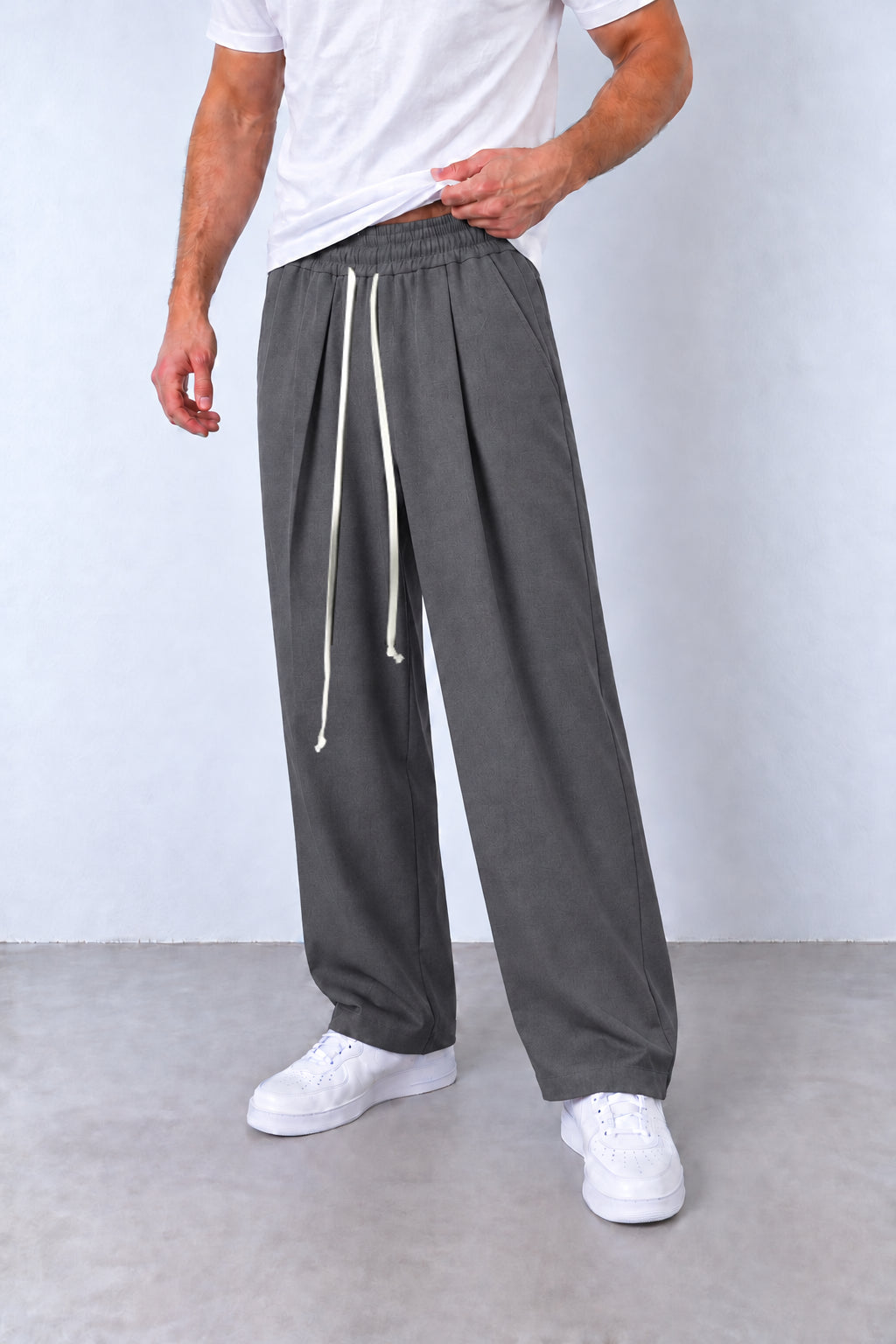 Pantaloni gri cu pense pe model, vedere din trei sferturi cu talie elastică și șnur lung.