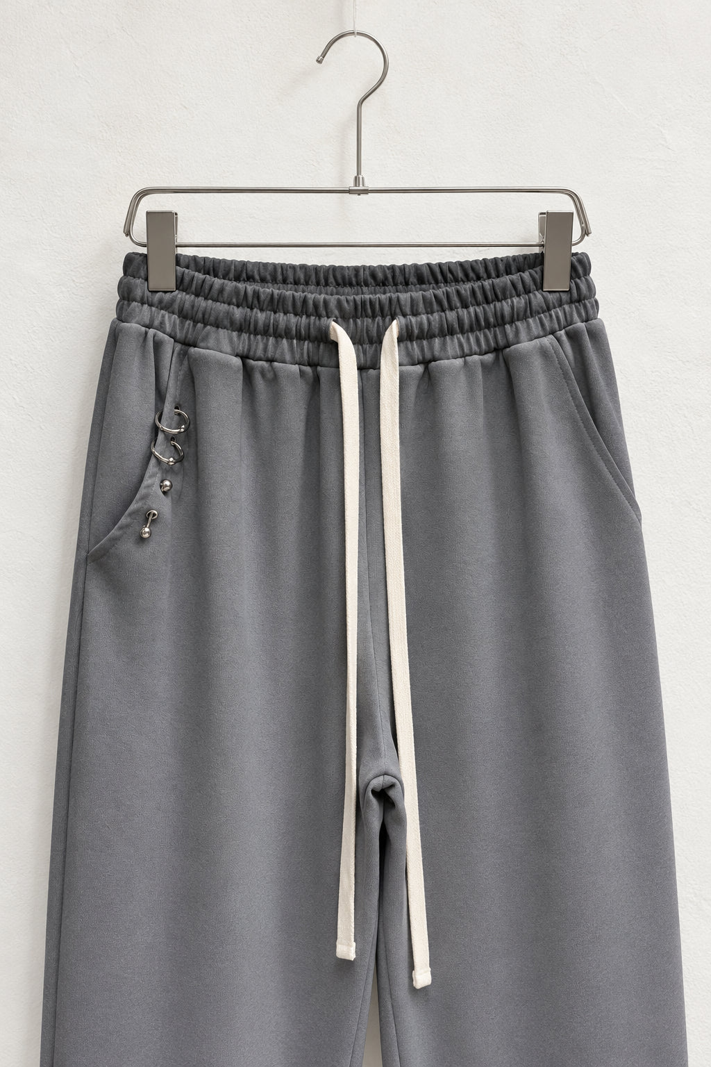 Detaliu talie elastică, șnururi plate și buzunar cu inele metalice la pantalonii gri.