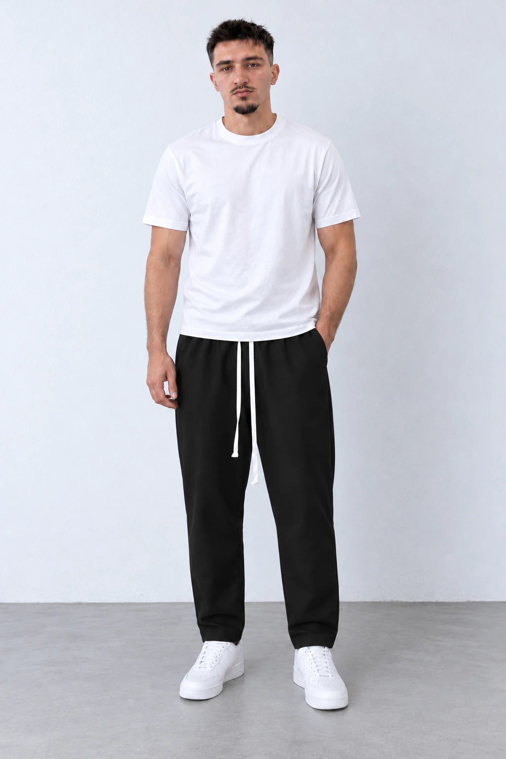 Pantaloni lungi negri carrot fit pentru bărbați, cu șnur alb lung, purtați cu tricou alb și sneakers, vedere integrală.