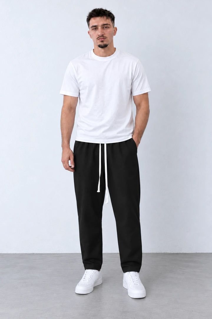 Pantaloni lungi negri carrot fit pentru bărbați, cu șnur alb lung, purtați cu tricou alb și sneakers, vedere integrală.