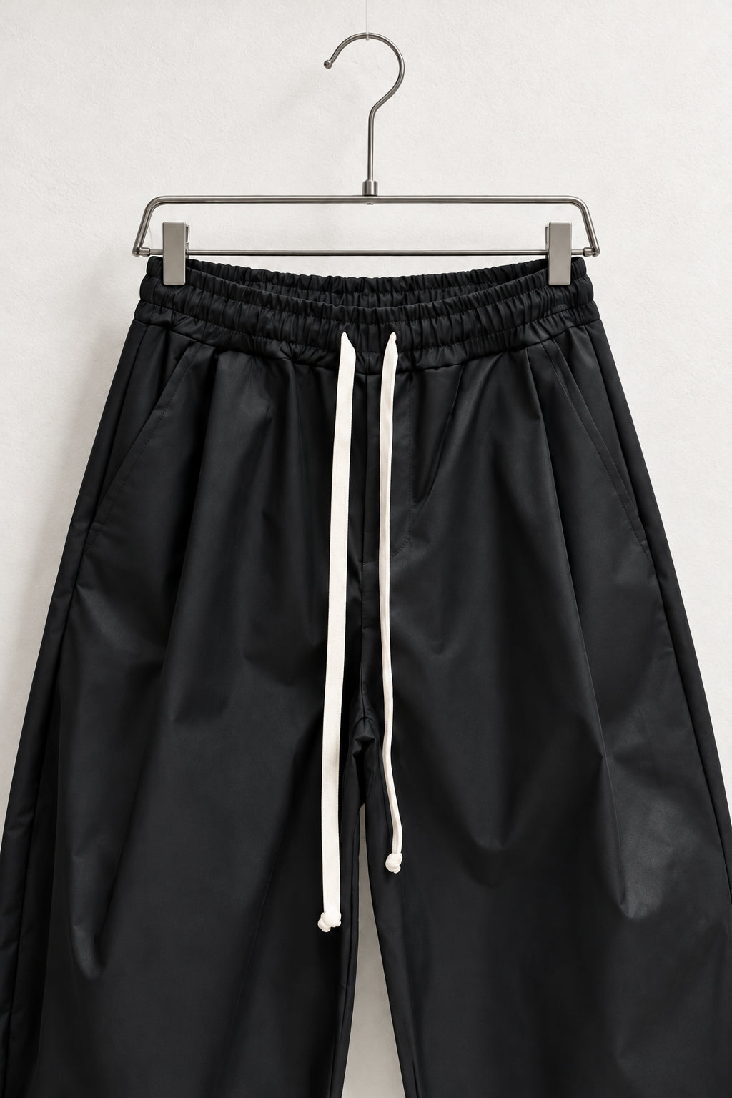 Detaliu talie elastică, buzunare și șnururi textile ale pantalonilor negri pe umeraș.