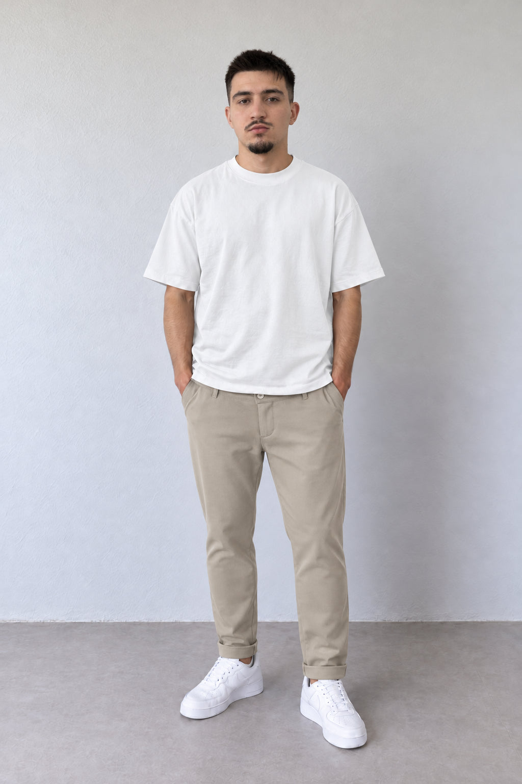Pantaloni bej slim fit pentru bărbați 008095-INV, cu talie clasică și tiv cu rever, purtați cu tricou alb și pantofi albi.