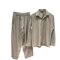 Un set de haine de vară, relaxat, format din cămașă lungă cu nasturi și pantaloni lungi asortați, în culoare camel deschis. Accentul este pus pe un șnur alb, foarte lung, vizibil, care atârnă din talia elastică a pantalonilor. Hainele sunt atârnate pe umerașe într-un suport din metal negru, pe fundalul unui perete de cărămidă albă texturată, cu o plantă verde în ghiveci în colțul din dreapta.