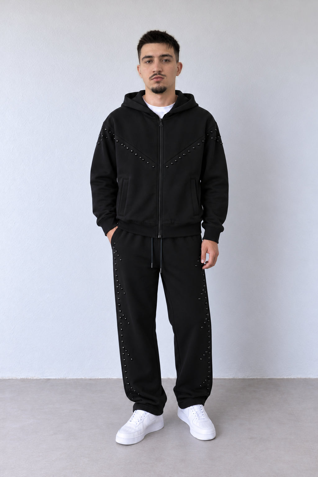 Trening negru pentru bărbați cu fermoar și aplicații tonale, ținută pe model în postură alternativă, cu croiala pantalonilor și aplicațiile vizibile.