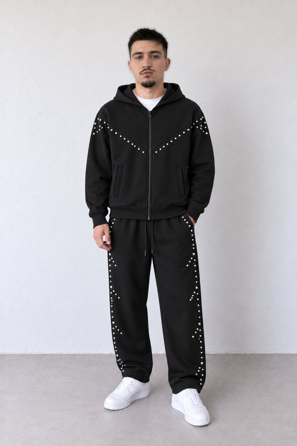 Trening negru pentru bărbați cu fermoar și capse argintii, ținută integrală pe model cu pantaloni asortați și sneakers albi.