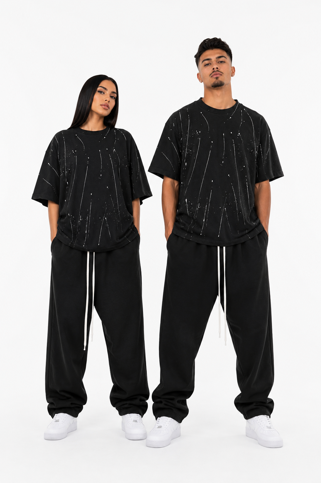 Model masculin și feminin purtând tricou negru oversized premium cu efect stropi de vopsea albă și pantaloni baggy streetwear negri, fotografie studio fashion luxury pe fundal alb.