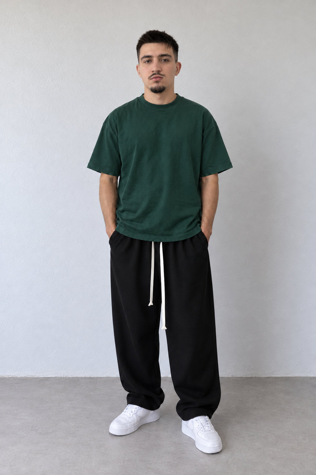 Tricou verde închis L815002 din bumbac, cu umeri căzuți și croială lejeră, purtat cu pantaloni negri și pantofi albi.